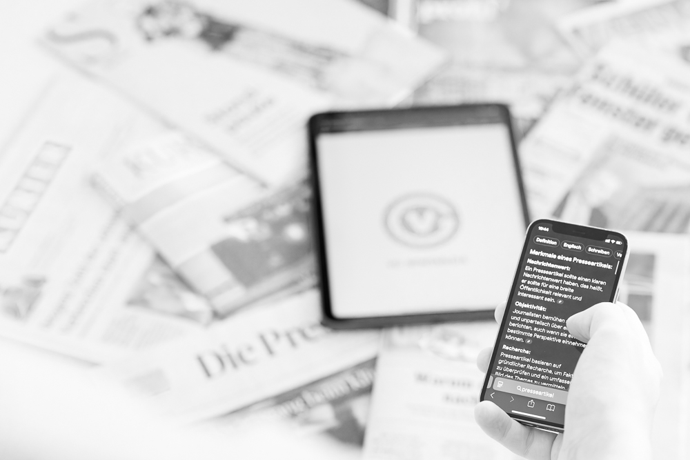 Eine symbolische Darstellung von Presseartikeln, die im Internet wiedergegeben werden. Zu sehen ist ein Tablet auf einem Stapel von Zeitungen und eine Hand, die auf einem Handy scrollt.